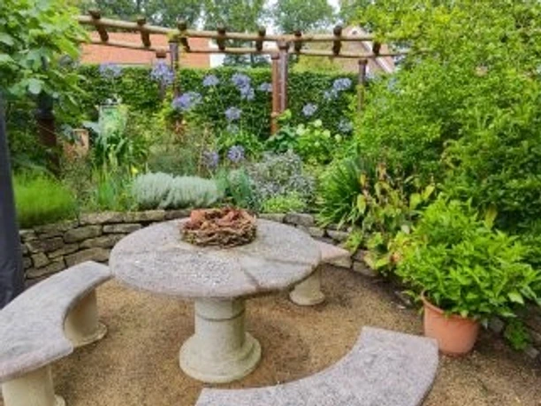 Sitzecke Garten Angelika und Holger Ihler.jpg Das Bild zeigt eine idyllische Gartensitzecke, die von üppigem Grün und blühenden Pflanzen umgeben ist. Ein runder Steintisch mit zwei halbmondförmigen Steinbänken wird von einem rustikalen Gartenzaun eingerahmt. Die farbenfrohe Blumenvielfalt und das satte Grün der Blätter schaffen eine friedliche und einladende Atmosphäre.