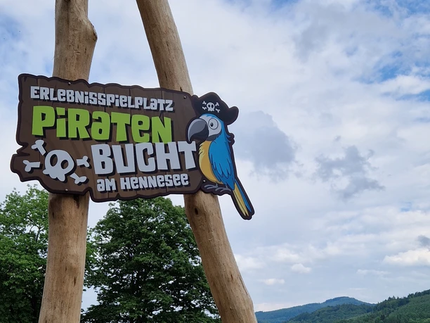 Logo Piratenbucht Hennesee Das Bild zeigt ein hölzernes Schild mit der Aufschrift "Erlebnisspielplatz Piratenbucht am Hennesee", darauf ein bunter Papagei.