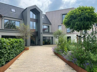 A modern building with large windows, surrounded by manicured greenery and a paved driveway.