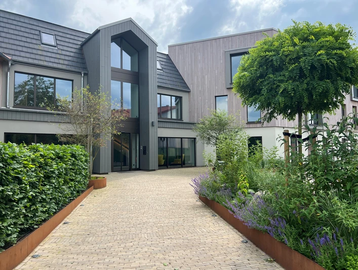 Thöles in Bücken Ein modernes Gebäude mit großen Fenstern, umgeben von gepflegtem Grün und einer gepflasterten Einfahrt.