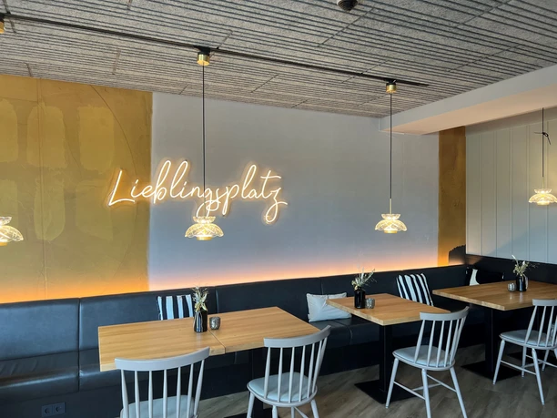 Cozy dining room with wooden tables, white chairs and neon "Lieblingsplatz" sign.