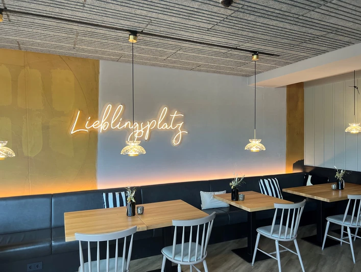 Gemütlicher Gastraum mit Holz-Tischen, weißen Stühlen und neonleuchtendem Schriftzug „Lieblingsplatz“.Cozy dining room with wooden tables, white chairs and neon "Lieblingsplatz" sign.Hyggelig spisestue med træborde, hvide stole og "Lieblingsplatz"-bogstaver i neon.Gezellige eetzaal met houten tafels, witte stoelen en neon "Lieblingsplatz" belettering.