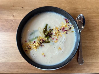 Eine cremige Spargelsuppe in einer dunklen Schüssel, dekoriert mit essbaren Blüten und Spargelstücken.A creamy asparagus soup in a dark bowl, decorated with edible flowers and pieces of asparagus.En cremet aspargessuppe i en mørk skål, dekoreret med spiselige blomster og aspargesstykker.Een romige aspergesoep in een donkere kom, versierd met eetbare bloemen en stukjes asperge.