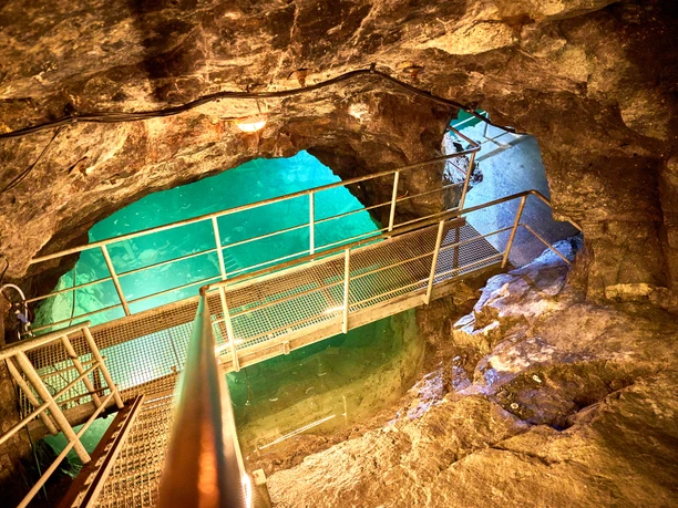 Metal bridge spans turquoise lake in natural underground space surrounded by rocks.
