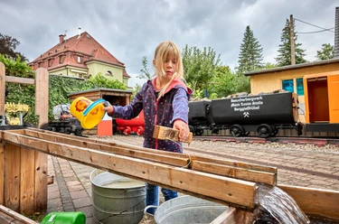 Child playing with water at the visitor mine in front of historic trolleys and a green landscape.