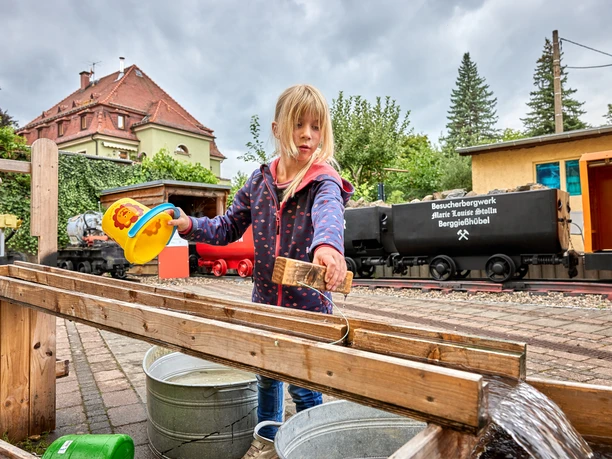 Kind speelt met water in de bezoekersmijn voor historische karretjes en een groen landschap.