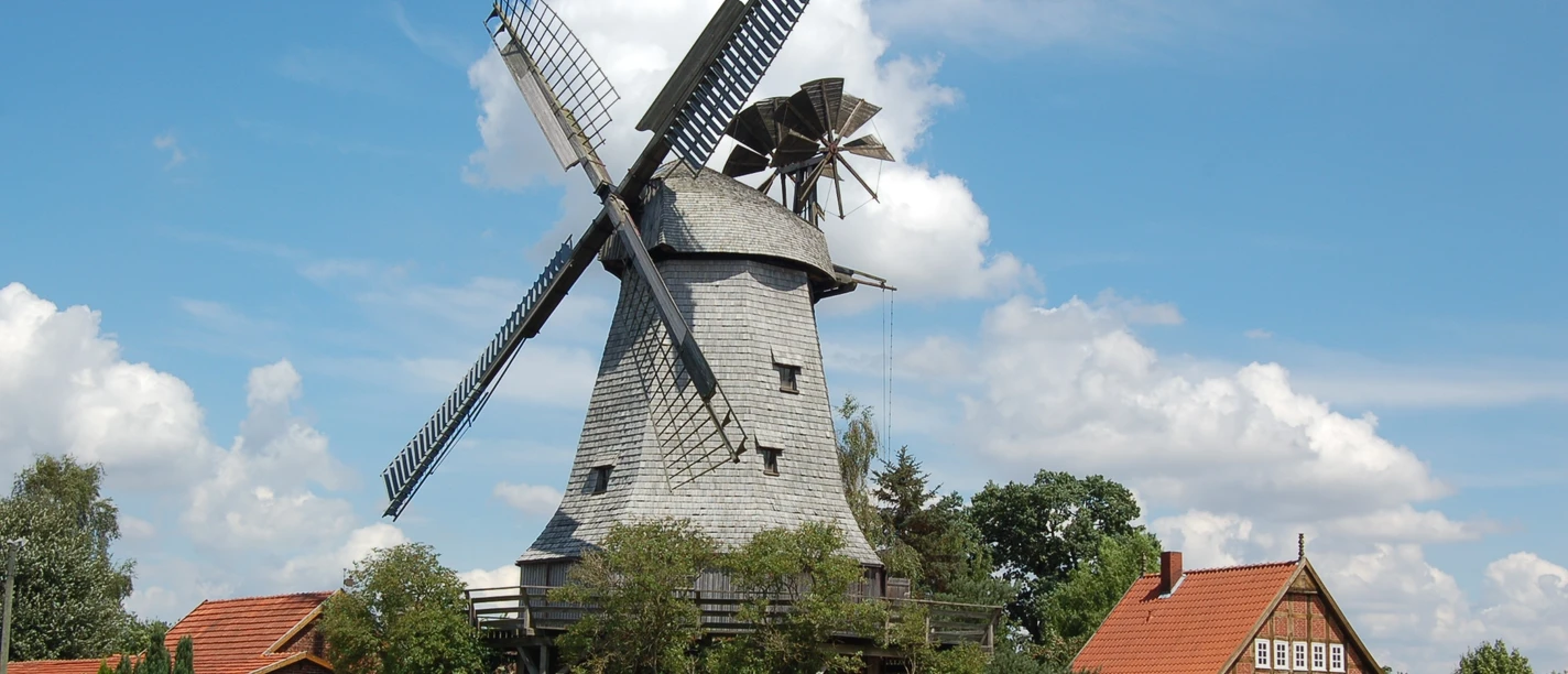 Eine historische Windmühle steht neben einem Backsteinhaus unter einem klaren blauen Sommerhimmel.