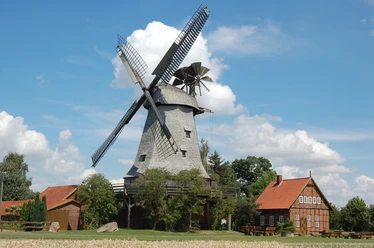 Stadt Petershagen_Meßlingen_Evelyn Hotze_Windmühle_Kornfeld im Sommer_quer.jpg Eine historische Windmühle steht neben einem Backsteinhaus unter einem klaren blauen Sommerhimmel.