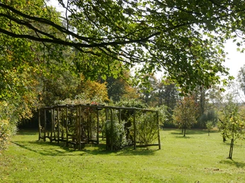 Wort-Weg HEGGE Ein hölzerner Pavillon inmitten einer grünen Wiese umgeben von Bäumen und blauen Himmel.
