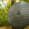 Wort-Weg HEGGE Metallscheibe mit eingraviertem Text im Wald, umgeben von Herbstblättern und Bäumen in Willebadessen.
