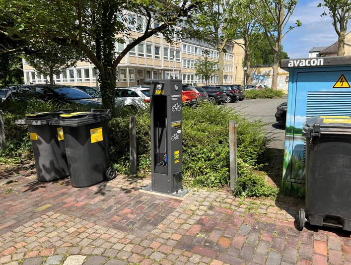 Graue ADAC-Service-Station für Fahrräder inmitten von Parkplatz, umgeben von Bäumen und Mülleimern.