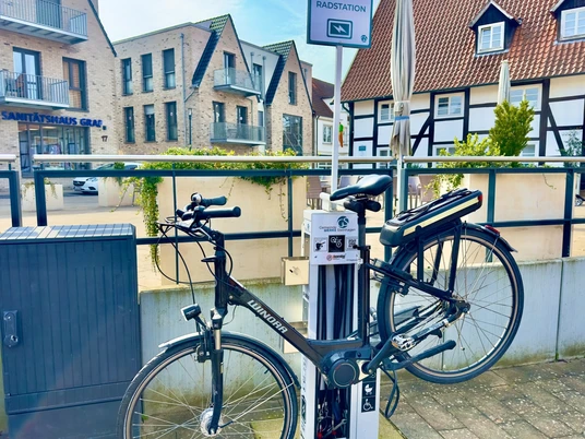 Fahrradstation mit aufgeladenem E-Bike in urbaner Umgebung, umgeben von modernen und traditionellen Gebäuden.