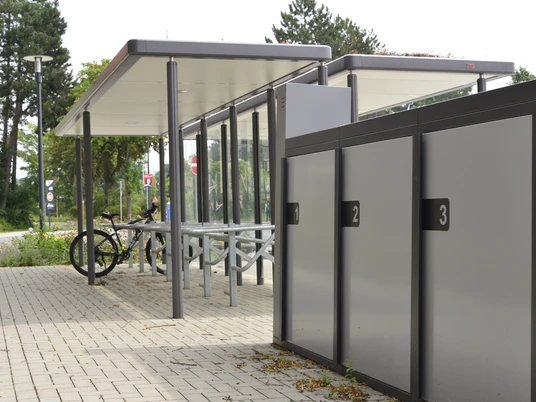 Mobilstation Steinhagen eBike Überdachte Fahrradständer und abschließbare Fahrradboxen auf einem gepflasterten Platz mit Bäumen.