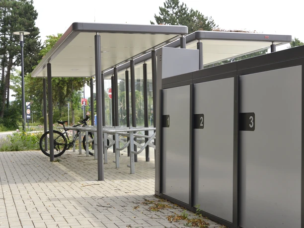 Mobilstation Steinhagen eBike Überdachte Fahrradständer und abschließbare Fahrradboxen auf einem gepflasterten Platz mit Bäumen.