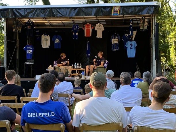 Arminia Podcast 1 - Copyright Gemeinde Steinhagen Open-Air-Bühne mit Zuschauern, zwei Menschen auf Stühlen, umgeben von Trikots und Bäumen im Hintergrund.