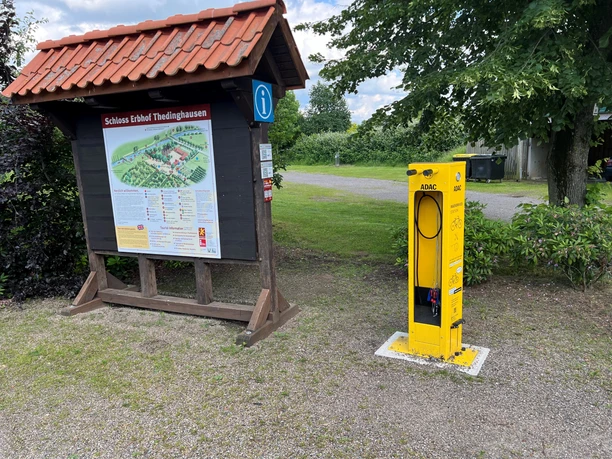 ADAC-Radservicestation neben Infotafel mit Schlosskarte; umgeben von Bäumen und Schotterweg.
