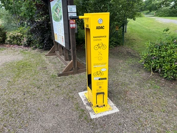 ADAC-Radservicestation Leuchtend gelbe ADAC-Radservicestation mit Werkzeugen und Luftpumpe, umgeben von grüner Natur.