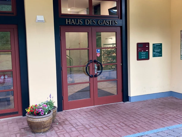Ingang Haus des Gastes Ingang van het "Haus des Gastes" met rode deuren, bloempot met kleurrijke bloemen aan de linkerkant, informatieborden aan de muur.