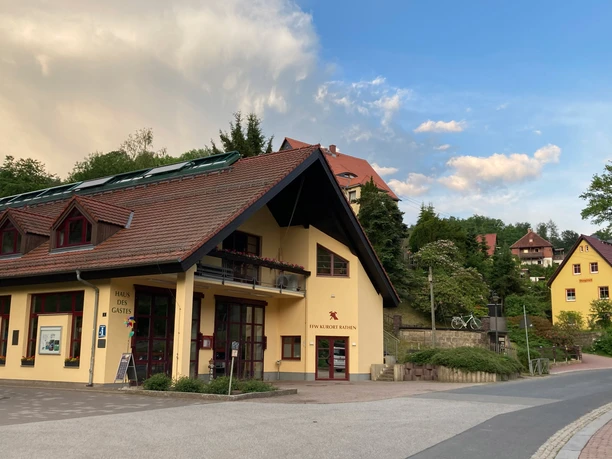 Dům pro hosty venku Žlutá budova s červenou střechou a solárními panely, obklopená zelenými stromy a dalšími domy, pod modrou oblohou s mraky.