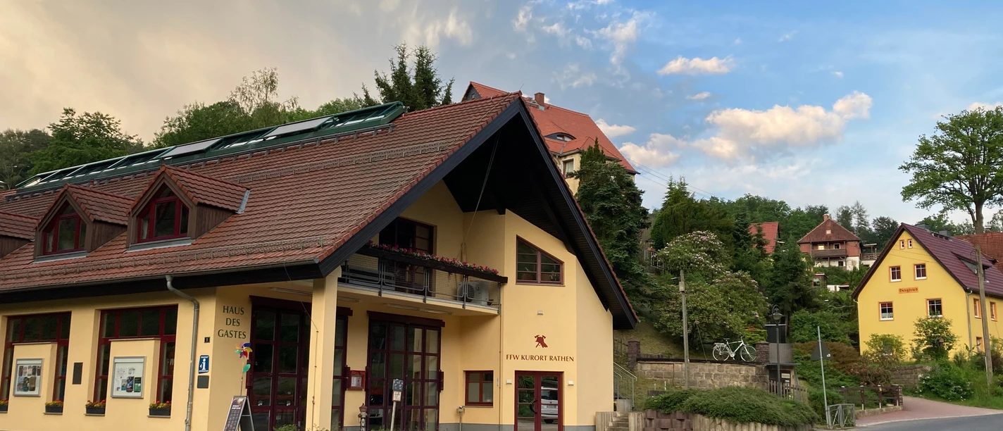 Haus des Gastes außen Gelbes Gebäude mit rotem Dach und Solarpanelen, umgeben von grünen Bäumen und weiteren Häusern, unter blauem Himmel mit Wolken.