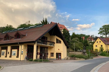 Huis van de gast buiten Geel gebouw met een rood dak en zonnepanelen, omringd door groene bomen en andere huizen, onder een blauwe lucht met wolken.
