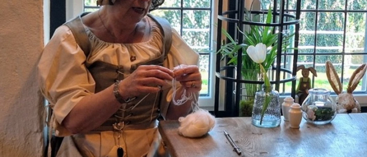 Geeske in´t Hörn bi´t Füür Frau in historischer Kleidung spinnt Wolle an Holztisch vor Fenster mit Frühlingsdekoration.