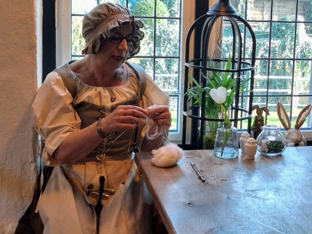 Geeske in´t Hörn bi´t Füür Frau in historischer Kleidung spinnt Wolle an Holztisch vor Fenster mit Frühlingsdekoration.