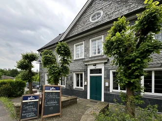 Landgasthof Zum Haus Olpe - Außenansicht Eine graue Fachwerkfassade mit grüner Tür und Schieferelementen, flankiert von Bäumen.