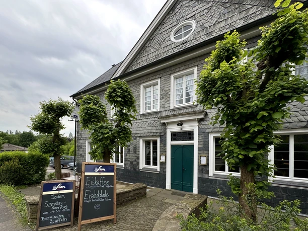 Landgasthof Zum Haus Olpe - Außenansicht Eine graue Fachwerkfassade mit grüner Tür und Schieferelementen, flankiert von Bäumen.