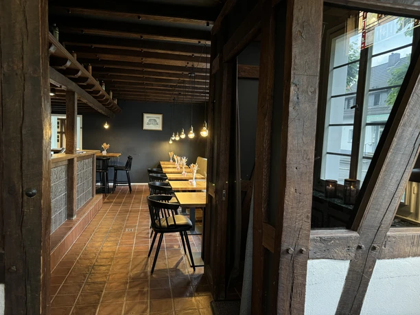Zum Haus Olpe - Ansicht Innenbereich Gemütliches Restaurant mit Holzbalken, langen Holztischen und warmem Licht von hängenden Lampen.