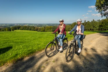 Heckberg Zwei Personen fahren lachend mit Fahrrädern auf einem ländlichen Weg durch eine grüne Landschaft.