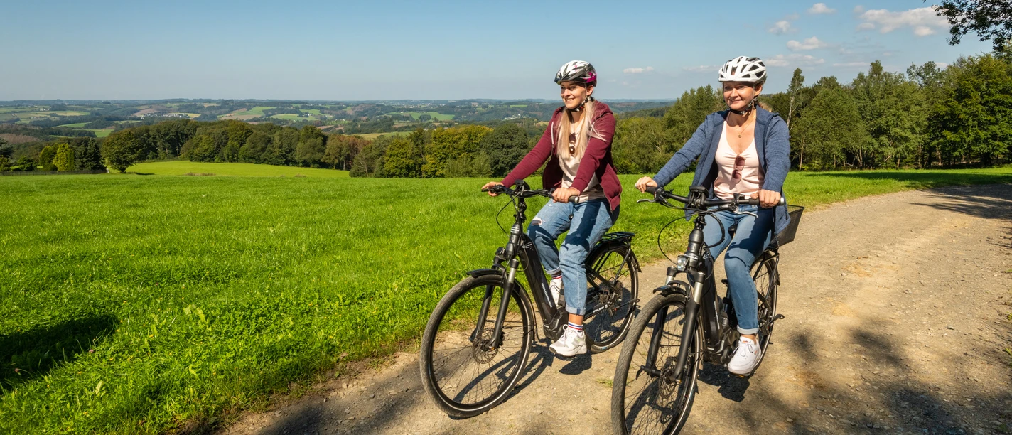 Heckberg Zwei Personen fahren lachend mit Fahrrädern auf einem ländlichen Weg durch eine grüne Landschaft.
