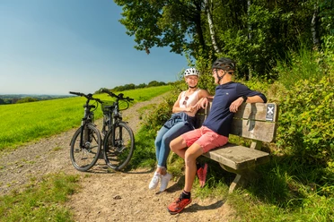 Heckberg Paar entspannt sich auf einer Bank am Radweg, mit zwei Fahrrädern und grüner Landschaft im Hintergrund.