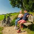 Heckberg Paar entspannt sich auf einer Bank am Radweg, mit zwei Fahrrädern und grüner Landschaft im Hintergrund.