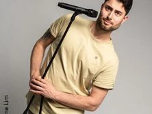 Ein humorvoller junger Mann in beiger Kleidung posiert mit Mikrofon in lockerer Haltung vor grauem Hintergrund.