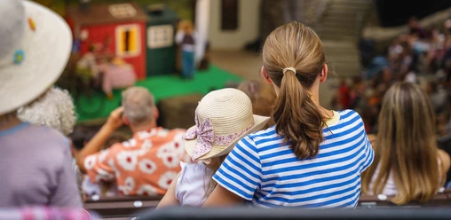 Familientag01(c)Lukas_Dahle Blick von hinten auf eine Frau, die mit ihrem Kind, das einen Hut trägt, auf einer Zuschauerbank sitzt und auf eine Bühne hinunterblickt.