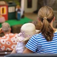 Familientag01(c)Lukas_Dahle Blick von hinten auf eine Frau, die mit ihrem Kind, das einen Hut trägt, auf einer Zuschauerbank sitzt und auf eine Bühne hinunterblickt.