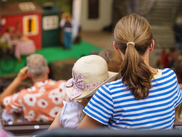 Familientag01(c)Lukas_Dahle Blick von hinten auf eine Frau, die mit ihrem Kind, das einen Hut trägt, auf einer Zuschauerbank sitzt und auf eine Bühne hinunterblickt.