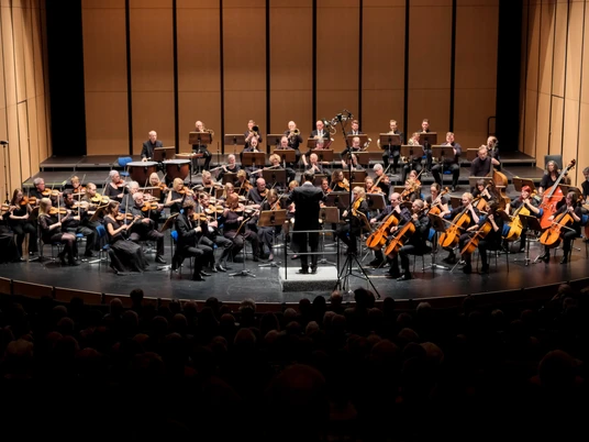 Sinfonieorchester Paderborn_Sinfoniekonzert-2024_Pressefoto 4.jpg Ein Sinfonieorchester spielt in einem Konzertsaal, dirigiert von einem schwarz gekleideten Dirigenten.