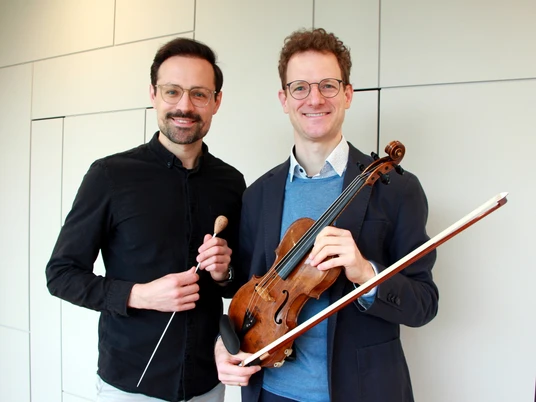 Sinfonieorchester Paderborn_Sinfoniekonzert-2024_Pressefoto 1.JPG Zwei Musiker mit Brillen halten Taktstock und Violine, lächelnd vor einer modernen, neutralen Wand.