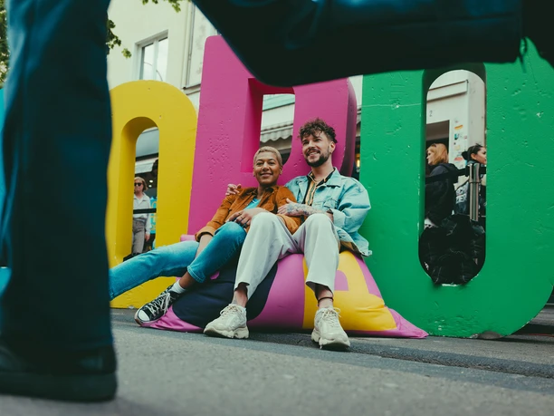 c/o pop in Cologne Two people sit relaxed on colorful beanbag cushions in front of large, colorful letters "c/o pop". In front of them, a person walks past the picture. The scene takes place on a street in Cologne, surrounded by passers-by and urban vibrancy.