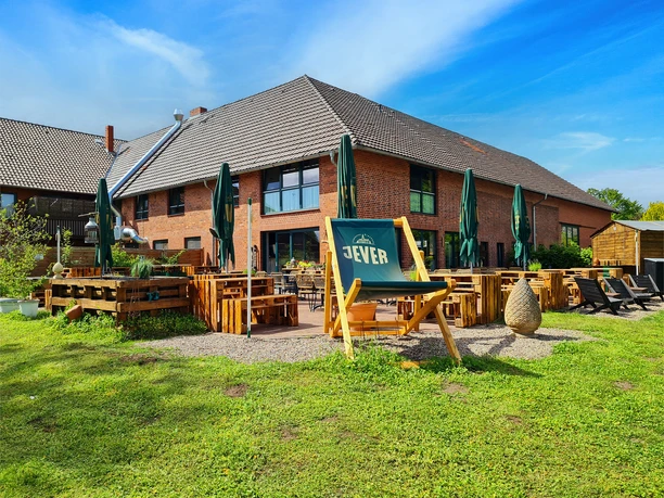 Kruegers_Terrasse_Biergarten_Außenansicht_Rückseite_Restaurant.jpg Großer Biergarten mit Sonnenschirmen und Liegestühlen, grüner Rasen im Vordergrund, hinterer Restaurantbereich.