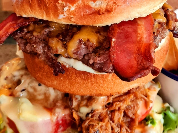 Kuegers_Triple_Burger.jpg Ein hoch aufgeschichteter Burger mit saftigem Fleisch, Speck und reichhaltigem Käse auf frischem Brioche.