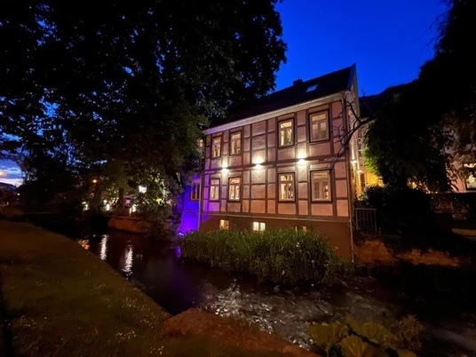 Pfälzer Weinstube 1.jpg Historisches Fachwerkhaus abends beleuchtet, neben einem Bach, umgeben von Bäumen.