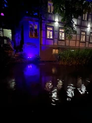 Pfälzer Weinstube 2.jpg Traditionelle Fachwerkfassade bei Nacht, durch lila Licht beleuchtet, spiegelt sich im Wasser.