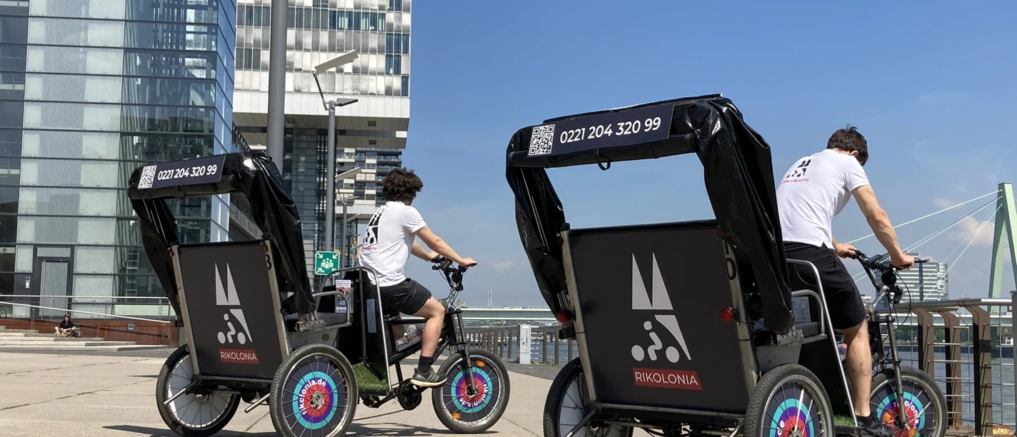 Rikolonia Citytour Rheinauhafen Two rickshaw drivers stand in Cologne's Rheinauhafen