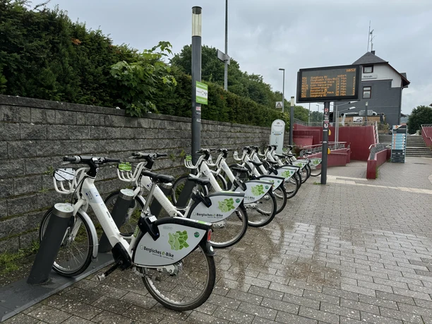 E-Bike Verleihstation am Bahnhof Rösrath Fahrradstation mit mehreren weißen Leihfahrrädern vor einem Bahnhof, umgeben von grünen Sträuchern.