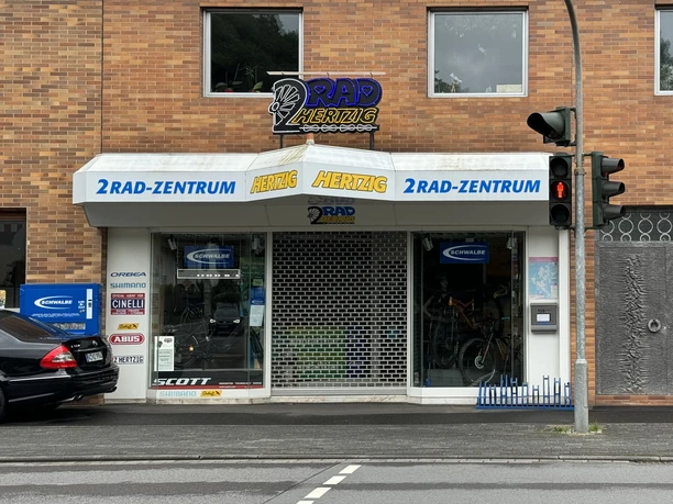 Zweiradzentrum Hertzig Fahrradgeschäft an belebter Straßenecke mit Radladen-Schild, Schaufenster und geparktem Auto davor.