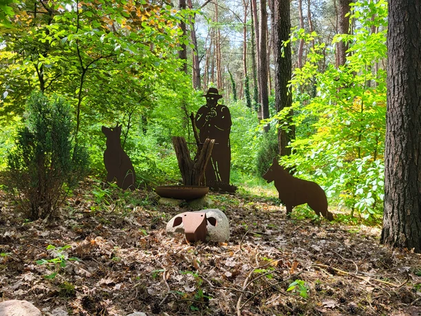 Infogarten Lorup_Book_Aufnahme TH ©Naturpark Hümmling (69).jpg Metallfiguren eines Schäfers mit Hunden und Schaf zwischen Bäumen im sonnendurchfluteten Wald.