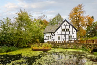 Emscherquellhof, Holzwickede <p>Fachwerkhaus am Teich mit grüner Umgebung, bewölkter Himmel, herbstliche Bäume im Hintergrund.</p>